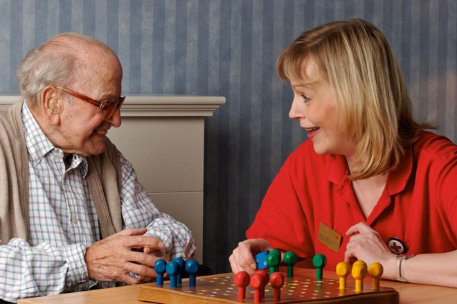 Bild zeigt eine DRK Mitarbeitende mit einem Senior an einem Tisch sitzend und ein Brettspiel (Mensch ärgere dich nicht) spielen