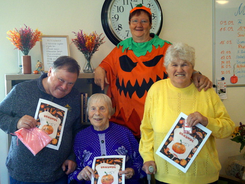 Bild zeigt drei Tagesgäste bei der Siegerehrung des Erntedankfestes. Jeder hält eine Urkunde mit der Platzierung und seinen Gewinn in der Hand. Es sind ein Mann und zwei Frauen. Im Hintergrund ist eine Mitarbeiterin in einem Kürbiskostüm zu sehen.