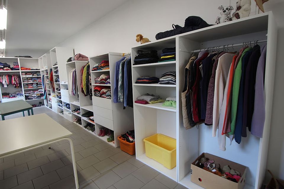 898f14fd-a31e-40a7-9c3b-5c97a5b0c8e8 Bild zeigt einen Blick in einen Raum der Kleiderkammer. Es sind lange offene Schrankreihen mit Fächern und Schubladen zu sehen in fast alle liegen bzw. hängen Kleidungsstücke und stehen Schuhe.