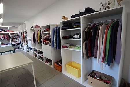 Bild zeigt einen Blick in einen Raum der Kleiderkammer. Es sind lange offene Schrankreihen mit Fächern und Schubladen zu sehen in fast alle liegen bzw. hängen Kleidungsstücke und stehen Schuhe.