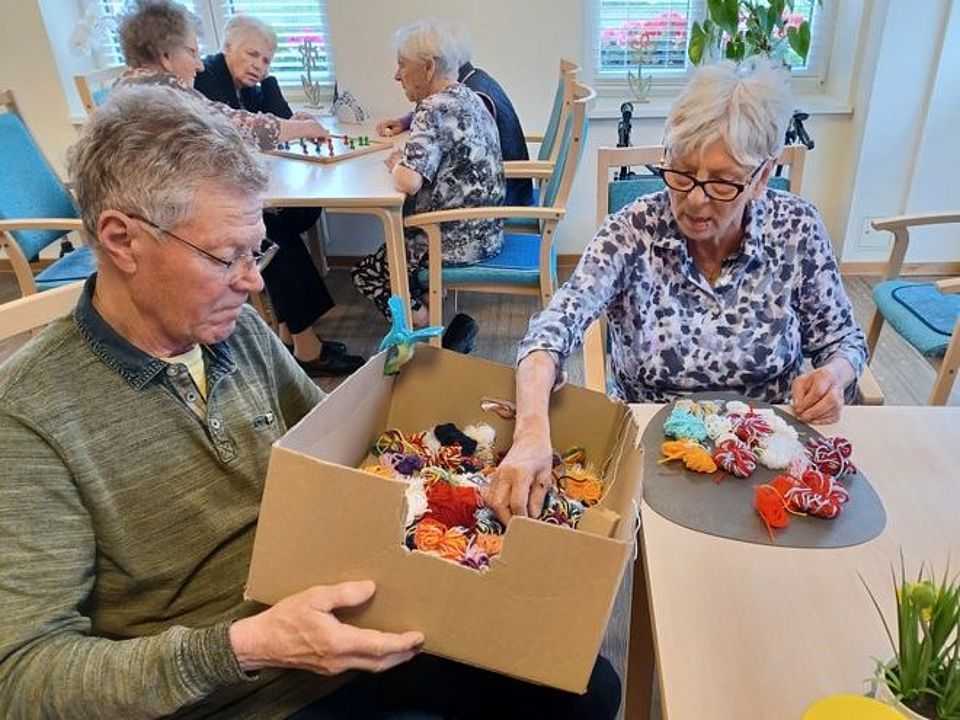 Bild zeigt den Aufenthaltsraum der Tagespflege. Es sind zwei Tische zu sehen. Am hinteren Tisch sitzen 4 Besucherinnen der Tagespflege und spielen "Mensch ärgere dich nicht". Am vorderen Tisch sitzen ein Besucher und eine Besucherin der Tagespflege und sortieren aus einem großen Karton Wollreste in vielen verschiedenen Farben für ein Beschäftigungsprojekt.