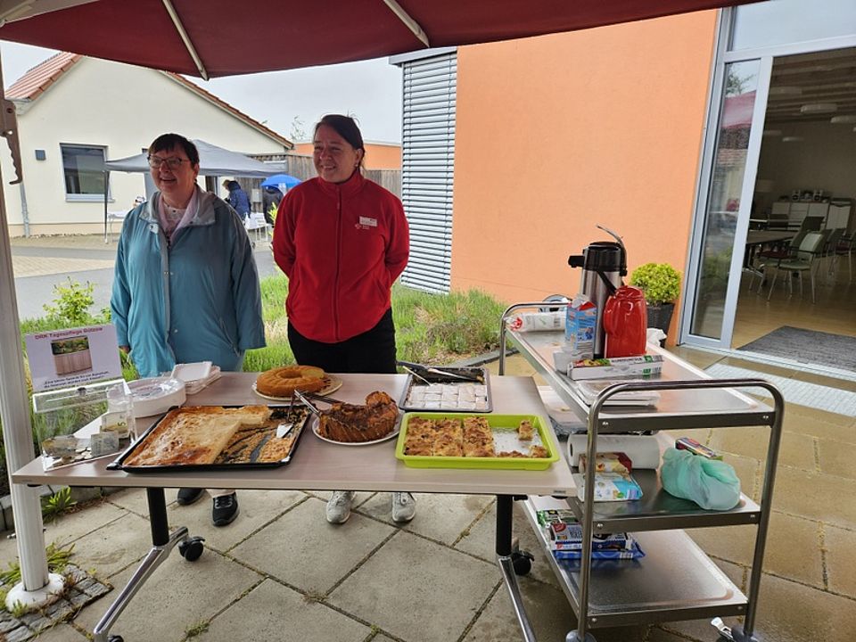 Bild zeigt zwei Mitarbeiterinnen der Tagespflege hinter einem Tisch stehen auf dem verschieden Kuchen zum Verkauf aufgebaut sind. Rechts neben dem Tisch steht ein Servierwagen mit Kaffee. Alles steht unter einem großem Sonnenschirm, da es leider regnet.