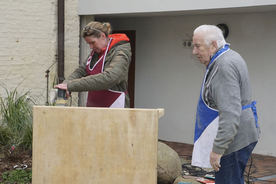 Bild zeigt: Eine jüngere Frau und ein älterer Mann arbeiten gemeinsam im Freien an einem Holzprojekt. Die Frau (links) trägt eine Jacke und eine dunkelrote Schürze und ist damit beschäftigt, mit einer Maschine (Schleifgerät) die Oberfläche einer großen Holzplatte zu bearbeiten. Der Mann (rechts) trägt eine blau-weiße Schürze, eine Strickjacke und Jeans und beobachtet die Arbeit. Die Szene zeigt eine gemeinsame handwerkliche Aktivität in der Tagespflege.