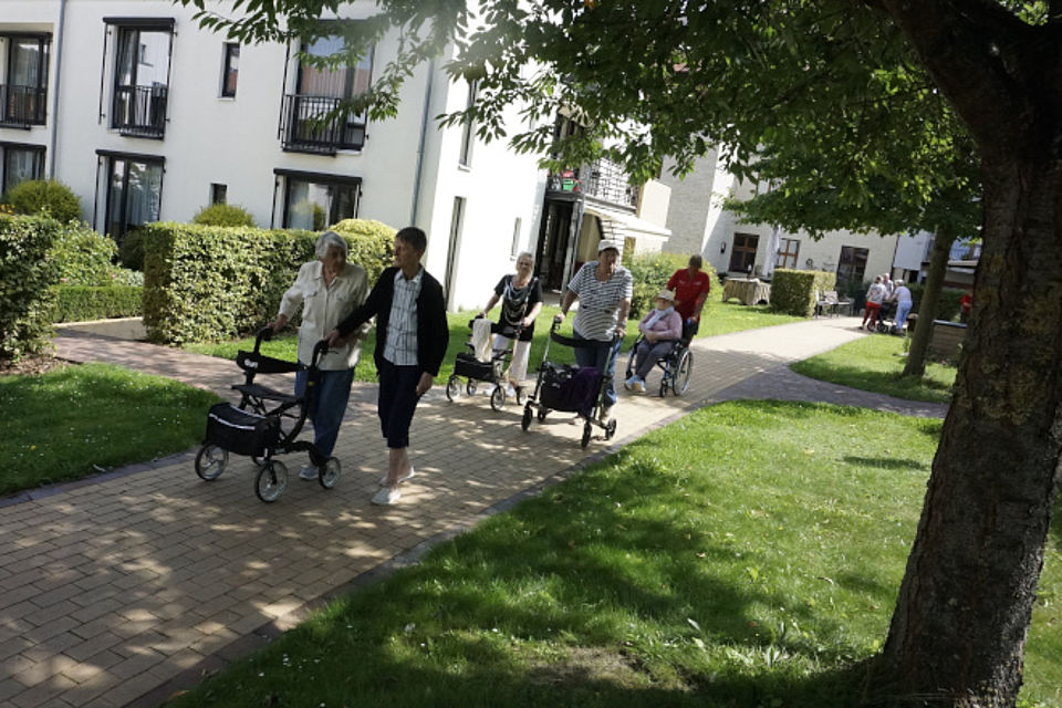 Bild zeigt: Eine Gruppe von Senioren macht einen Spaziergang mit Rollatoren und einem Rollstuhl auf einem gepflasterten Weg in der Außenanlage einer Wohn- und Pflegeeinrichtung. Eine Mitarbeiterin begleitet die Gruppe. Die Szene zeigt die Mobilität und Aktivierung der Tagesgäste bei einem gemeinsamen Spaziergang im Grünen, umgeben von gepflegten Hecken, Bäumen und Rasenflächen.