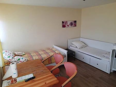 Bild zeigt einen Blick in ein Zimmer im Frauenschutzhaus, eingerichtet mit zwei Betten, eines davon als Tagessofa nutzbar, weiterhin Tisch und Stühle