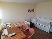 Bild zeigt einen Blick in ein Zimmer im Frauenschutzhaus, eingerichtet mit zwei Betten, eines davon als Tagessofa nutzbar, weiterhin Tisch und Stühle
