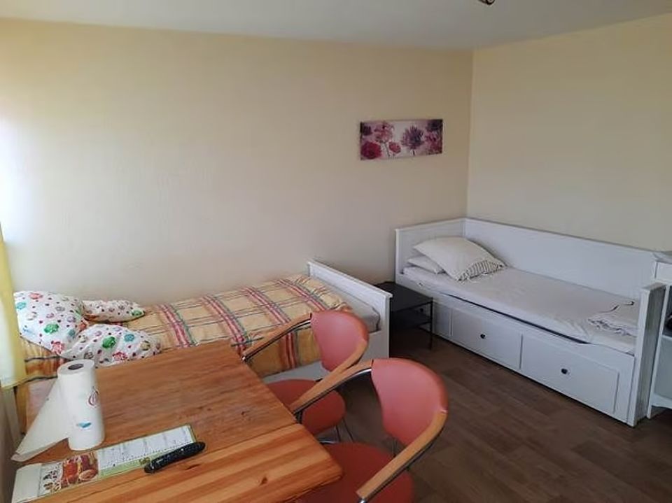 Bild zeigt einen Blick in ein Zimmer im Frauenschutzhaus, eingerichtet mit zwei Betten, eines davon als Tagessofa nutzbar, weiterhin Tisch und Stühle