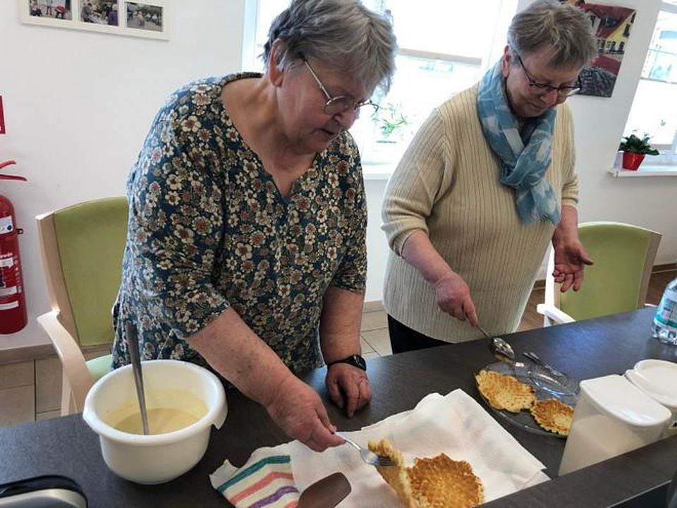 Bild zeigt zwei Besucherinnen der Tagespflege vor einem Tisch stehend. Sie backen Waffeln.