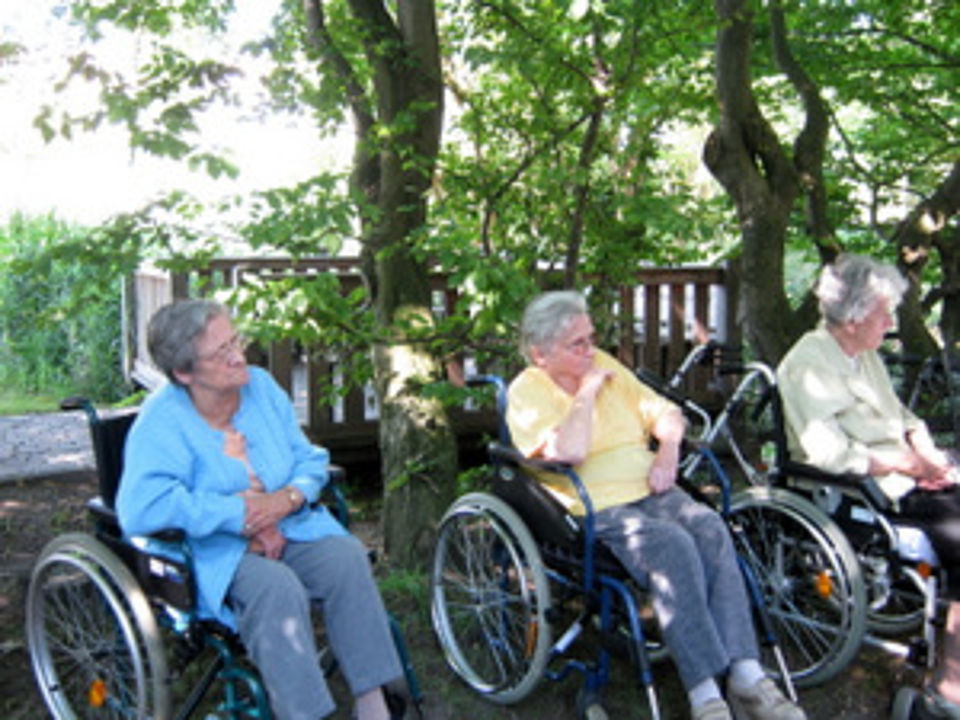 Bild zeigt eine Gruppe von Bewohnern, sitzend mit Rollstuhl im Park unter Bäumen