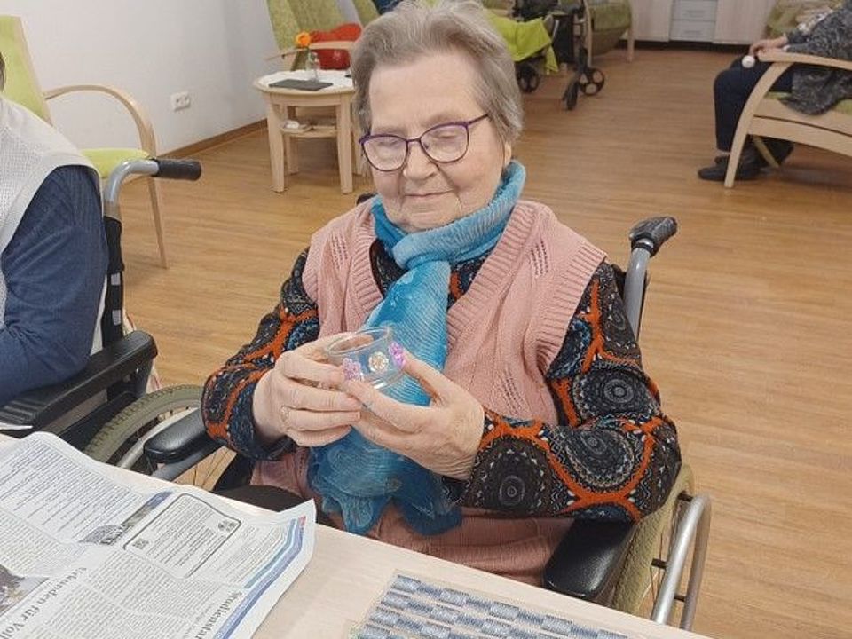 Bild zeigt Tagesgast Frau Hartwig in ihrem Rollstuhl am Tisch sitzend. In den Händen hält sie ein kleines gläsernes Windlicht, welches sie schon mit mehreren Motivstickern beklebt hat. Sie begutachtet ihr Werk und ist vom Gesichtsausdruck her sichtlich zu frieden.