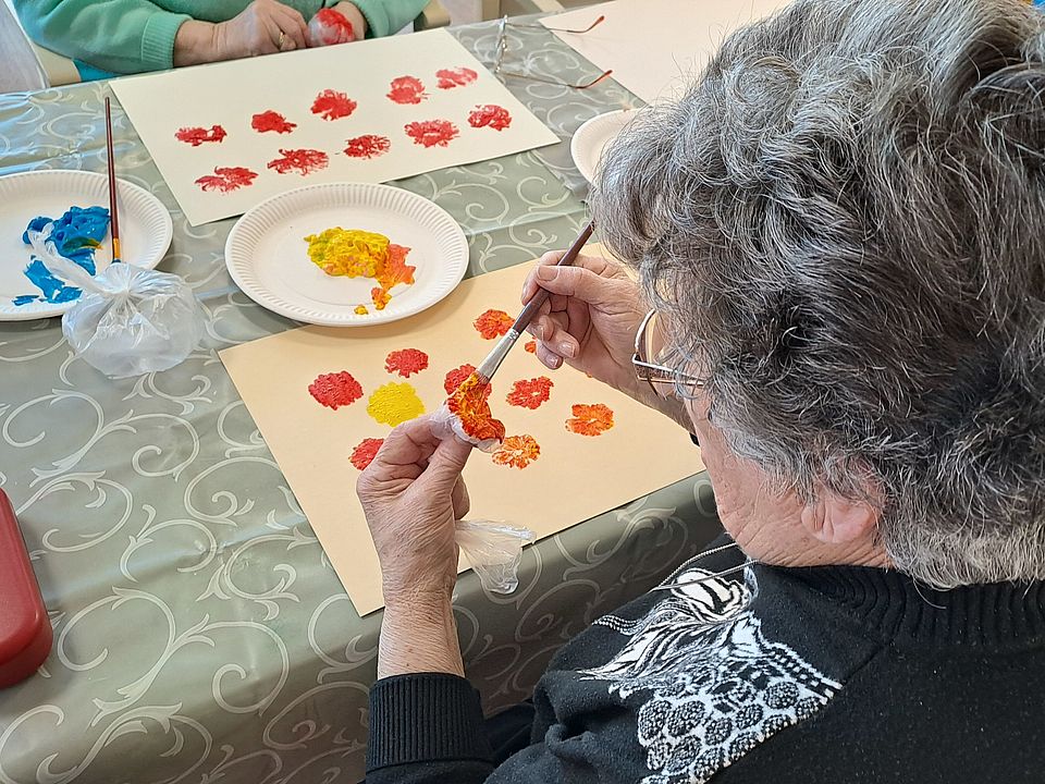 Bild zeigt Nahaufnahme einer Seniorin, die an einem Tisch sitzt und mit einem Pinsel gelbe und orangefarbene Tupfer auf ein beigefarbenes Papier aufträgt. Auf dem Tisch liegen weitere fertige Tupf-Muster in Rot- und Gelbtönen, eine Palette mit Farbtupfern sowie Farbteller mit leuchtender roter und blauer Farbe.