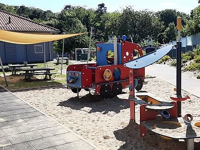 Bild zeigt den Spielplatz, mit Geräten die in einem Sandfeld stehen. Im Hintergrund ist ein Sitzplatz mit Sonnensegel zu sehen.