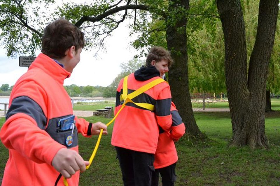 Rettungsmitteltest Bild zeigt die drei in der Bildbeschreibung genannten Personen beim Test der Rettungsleine. Diese Leine ist um die Schultern und Körper einer Person gebunden und diese Person hält eine weitere Person im Rettungsgriff. Die dritte Person mit dem Ende der Leine in der Hand unterstützt das an Land holen der anderen beiden Personen. Das ganze Szenario wird allerdings an Land, auf der Wiese gezeigt. Normalerweise findet die Leine bei einer Wasserrettung ihren Einsatz.