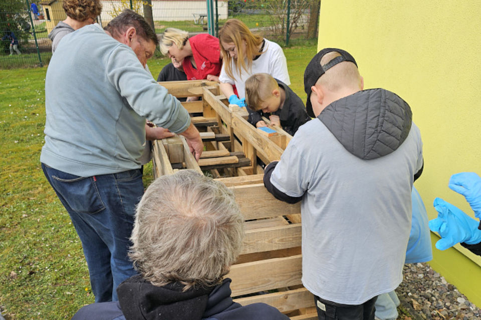 Bild zeigt: Eine Gruppe von Menschen unterschiedlichen Alters, darunter Senioren, jüngere Erwachsene und Kinder, arbeitet gemeinsam im Freien am Projekt Hochbeet. Sie sind um Holzpaletten und eine Konstruktion aus Latten versammelt und helfen beim Bau. Die Szene zeigt ein generationsübergreifendes Projekt ("Jung & Alt"), bei dem gemeinsam handwerkliche Fähigkeiten eingesetzt werden, zum Bau eines Hochbeets.