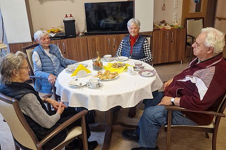 Bild zeigt: Vier Senioren sitzen um einen runden Tisch mit weißer Decke, auf dem Kaffeegeschirr und Gebäck stehen. Sie unterhalten sich in einem gemütlichen Raum, der im Hintergrund braune Schränke und einen Fernseher zeigt."