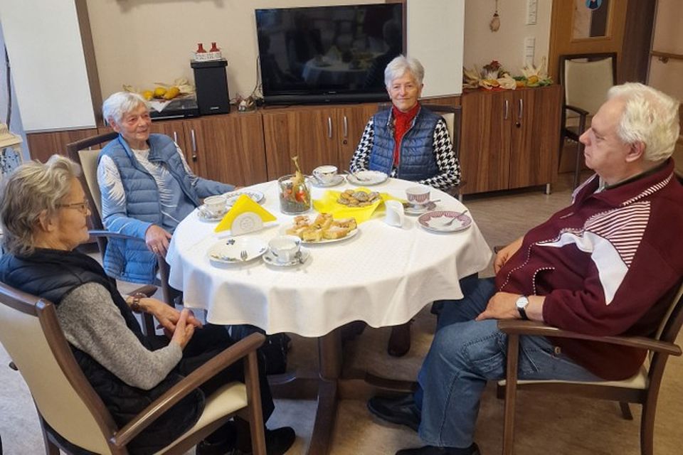 Kaffeerunde Bild zeigt: Vier Senioren sitzen um einen runden Tisch mit weißer Decke, auf dem Kaffeegeschirr und Gebäck stehen. Sie unterhalten sich in einem gemütlichen Raum, der im Hintergrund braune Schränke und einen Fernseher zeigt."