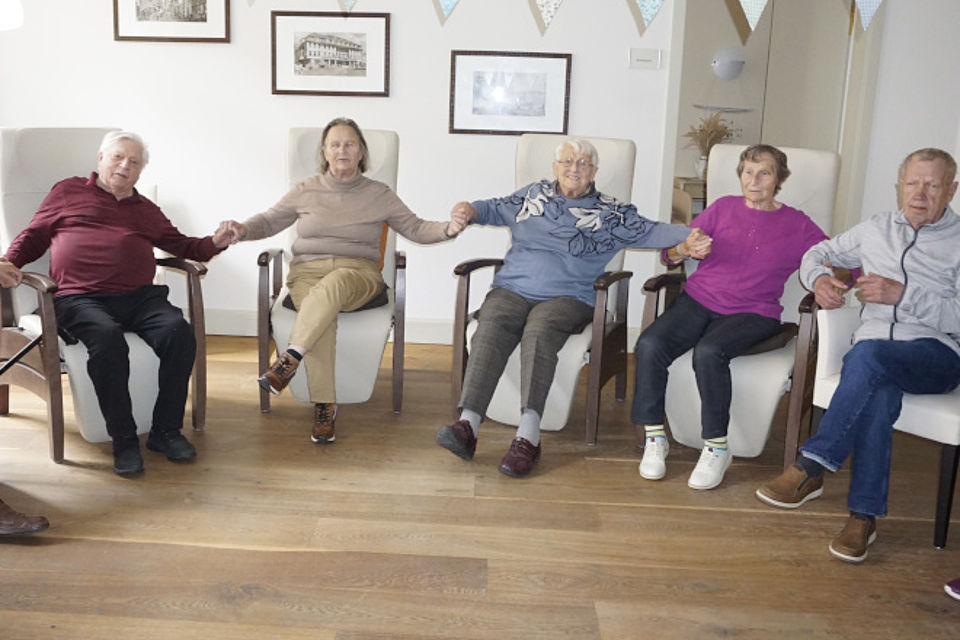 Bild zeigt: Fünf Senioren sitzen in bequemen Sesseln in einem Gemeinschaftsraum und halten sich an den Händen, wodurch eine Kette entsteht. Die vier Personen in der Mitte schauen freundlich in die Kamera, während der Mann ganz rechts etwas in seiner Hand hält und zur Seite blickt. Die Szene zeigt eine Bewegungsübung im Sitzen, die Gemeinschaft und Zusammenhalt in der Tagespflege  fördert. Im Hintergrund hängen Bilder an der Wand und eine Wimpelkette ziert den oberen Bereich.