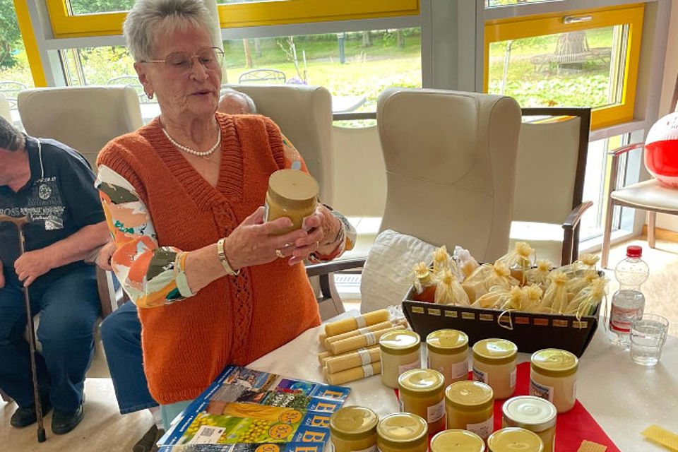 Bild zeigt: Eine ältere Dame, Ursula Wende, steht an einem Tisch und hält ein großes Glas mit Honig in ihren Händen, das sie interessiert betrachtet. Sie trägt eine orangefarbene Strickweste und eine Perlenkette. Auf dem Tisch vor ihr sind zahlreiche weitere Honiggläser, Honigbonbons oder -kerzen, die in Tüten verpackt sind, sowie ein Magazin oder eine Broschüre mit einem gelben Titel (möglicherweise über Bienen oder Honig) zu sehen. Die Szene findet im Rahmen des Mottotags "Fleißiges Bienchen" statt.