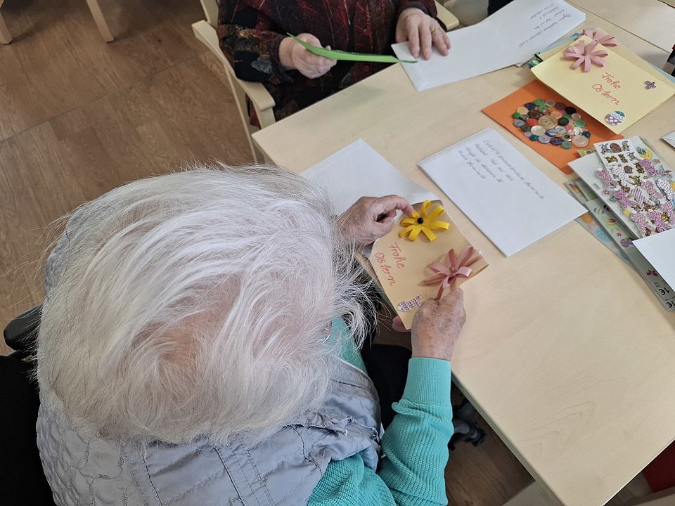 Bild zeigt: Aufsicht aus der Vogelperspektive auf eine Seniorin mit weißem Haar, die an einem hellen Holztisch eine selbstgebastelte Osterkarte verziert. Sie hält eine braune Karte in den Händen, auf der 'Frohe Ostern' steht und die mit einer gelben und einer rosafarbenen Papierblume geschmückt ist. Auf dem Tisch liegen weitere fertige, dekorierte Karten, beschriftete Briefumschläge und Bastelmaterialien wie bunte Knöpfe und Aufkleber für das Projekt 'Post mit Herz'.