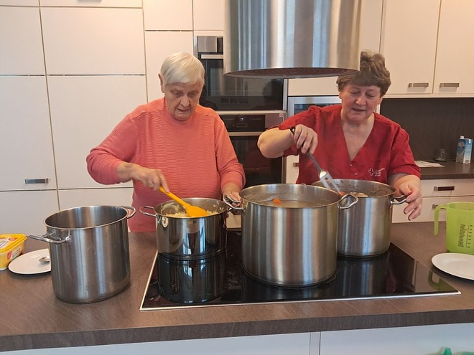 Bild zeigt eine Besucherin der Tagespflege, die zusammen mit einer Mitarbeiterin an der großen Kochinsel steht und in zwei sehr großen Töpfen Eisbein kochen