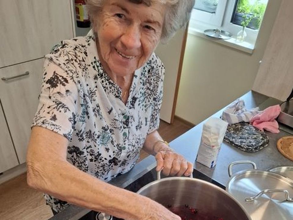 Bild zeigt eine ältere Frau am Herd stehnd und in einem großen Topf mit verschiedenen roten und dunkleblauen Beeren rühren. Neben ihr steht eine Tüte Zucker und ein weiterer Topf, der aber mit einem Deckel abgedeckt ist.
