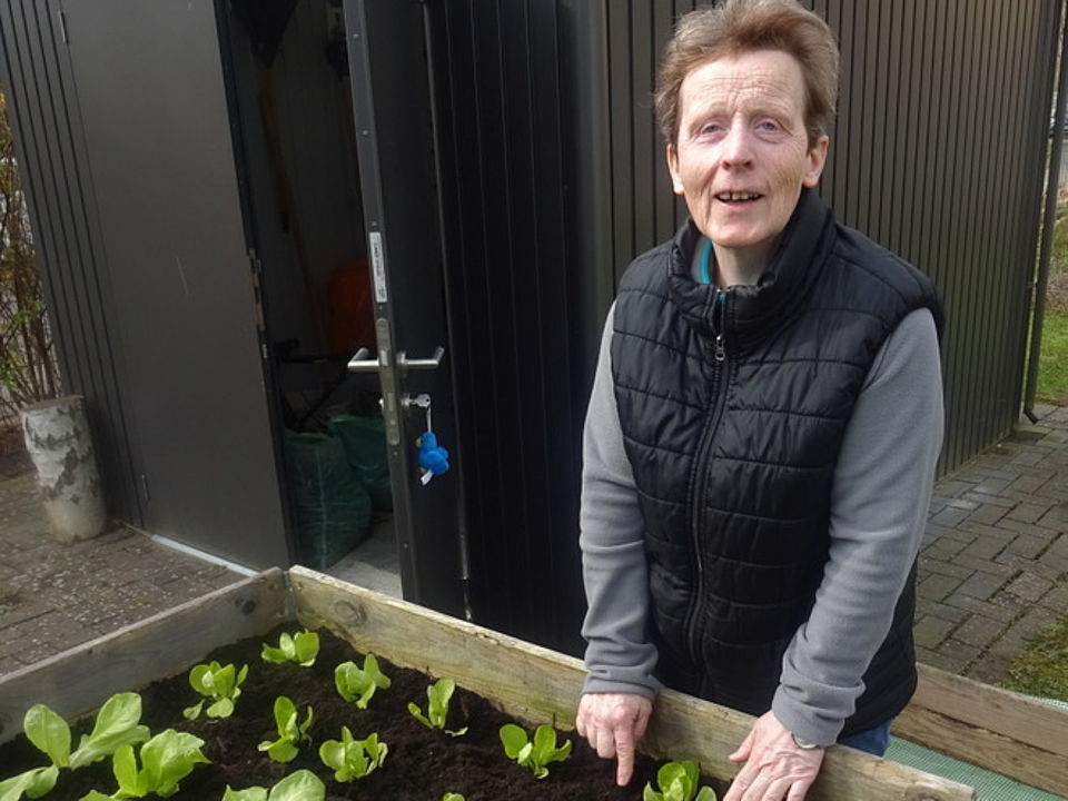 Bild zeigt Frau Ute Eislöffel vor dem zweiten Hochbeet stehend. Sie trägt eine graue Fliesjacke und darüber eine schwarze Steppweste. Vor ihr im Hochbeet sieht man ausgerichtete und eingepflanzte junge Salatpflanzen.