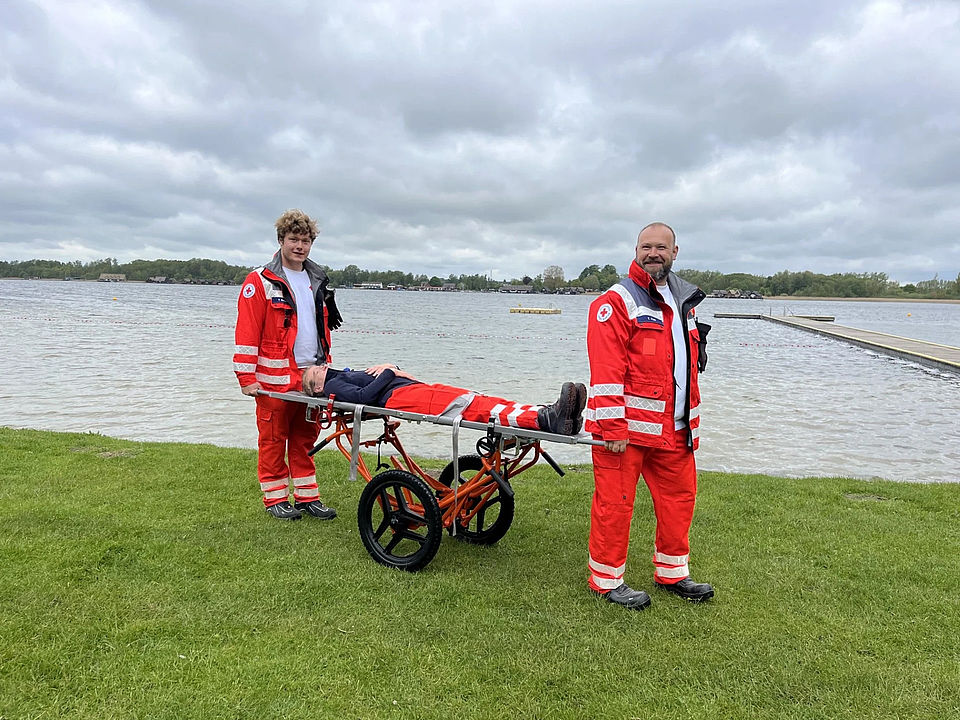 Die Rolltrage Bild zeigt zwei Personen in DRK-Einsatzkleidung, die mit einer fahrbaren Trage eine weitere Person transportieren. Im Hintergrund ist der Inselsee zu sehen.