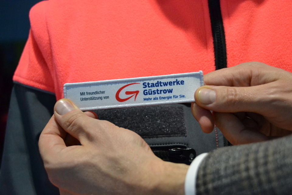 Juniorretter Bild 4 Bild zeigt in Nahaufnahme den Sponsoring-Patch mit der Aufschrift: Mit freundlicher Unterstützung von dann kommt das Logo der Stadtwerke. im Hintergrund ist die Jacke von Hans-Erik zu sehen mit der Stelle, an welcher der Patch nun gleich angebracht wird