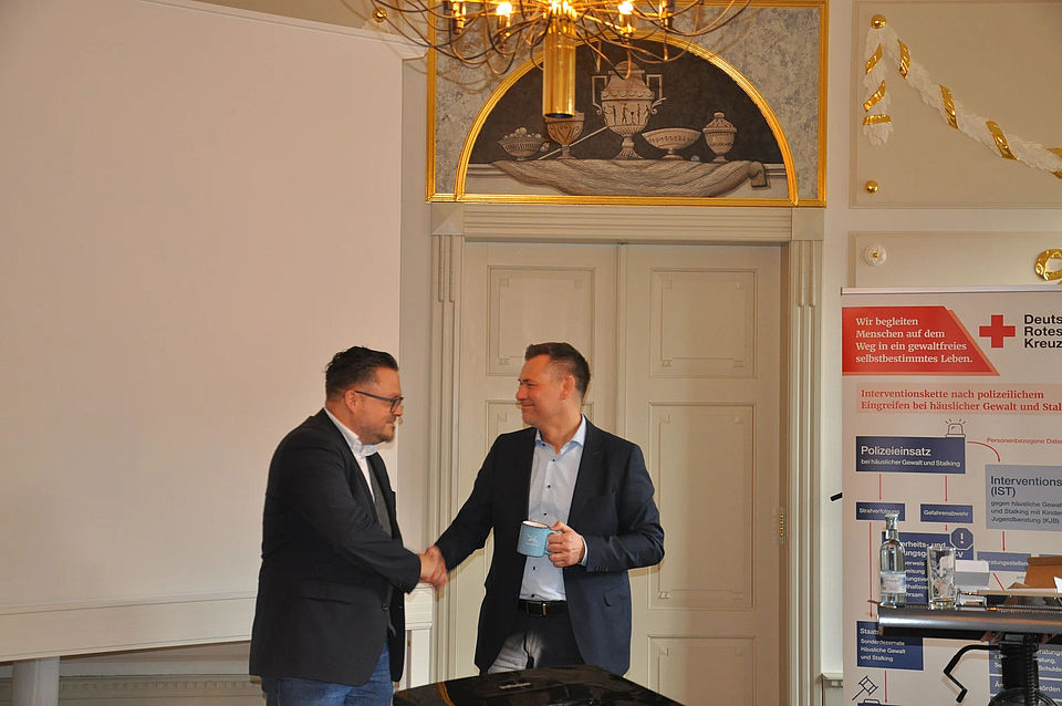 Bild zeigt Bürgermeister Sascha Zimmermann bei der Gratulation von Alexander Plass, Vorstandsvorsitzender des DRK-Kreisverbandes Güstrow e.V. Sie schütteln die Hände und Bürgermeister Zimmermann hält eine Bechertasse in himmelblau mit einem Spruch darauf in der linken Hand. Den Spruch kann man leider nicht lesen.