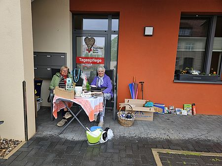 Bild zeigt zwei Frauen hinter einem Klapptisch sitzend. Auf dem Tisch liegen und stehen mehrere Artikel, welche verkauft werden sollen. Am Boden erkennt man, dass es regnet.
