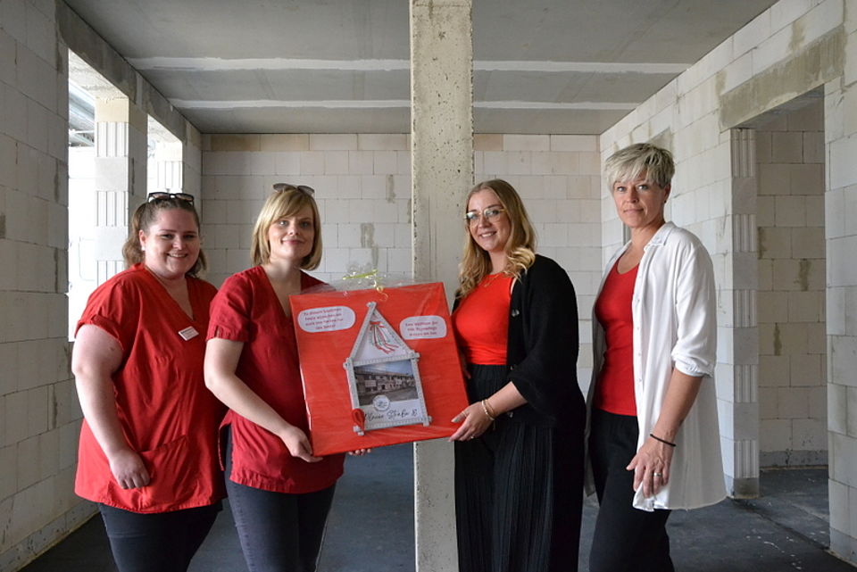 Bild zeigt Laura Holtz und Katharina Möbius mit einem selbstgebasteltes Geschenk. Eine Leinwand mit aus einem Zollstock erstelltem Haus und Bild vom Bau und guten Wünschen. Neben ihnen stehen Frau Lea Schulz und Stefanie Kelch.
