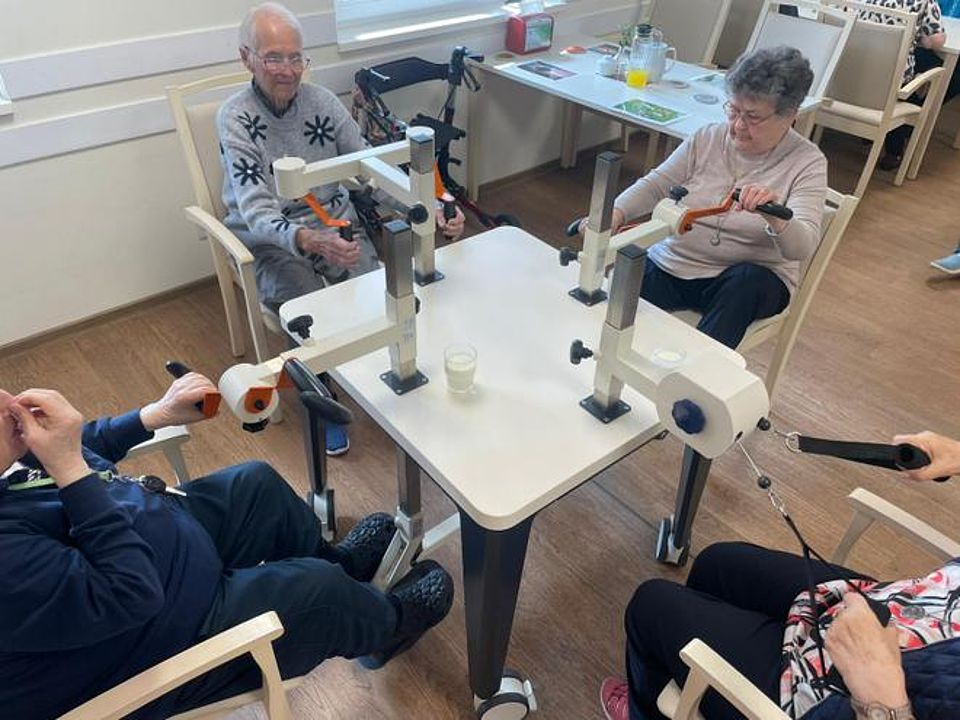 Bild zeigt einen Tisch an dem verschiedene Trainingsgeräte (2 Handpedale, 1 Art Butterfly für Hände und Oberarme und 1 Zuggerät ähnlich einer Rudermaschine) An dem Tisch sitzen 4 Tagesgäste Jeder vor einem Gerät und betätigen sich und erzählen dabei.