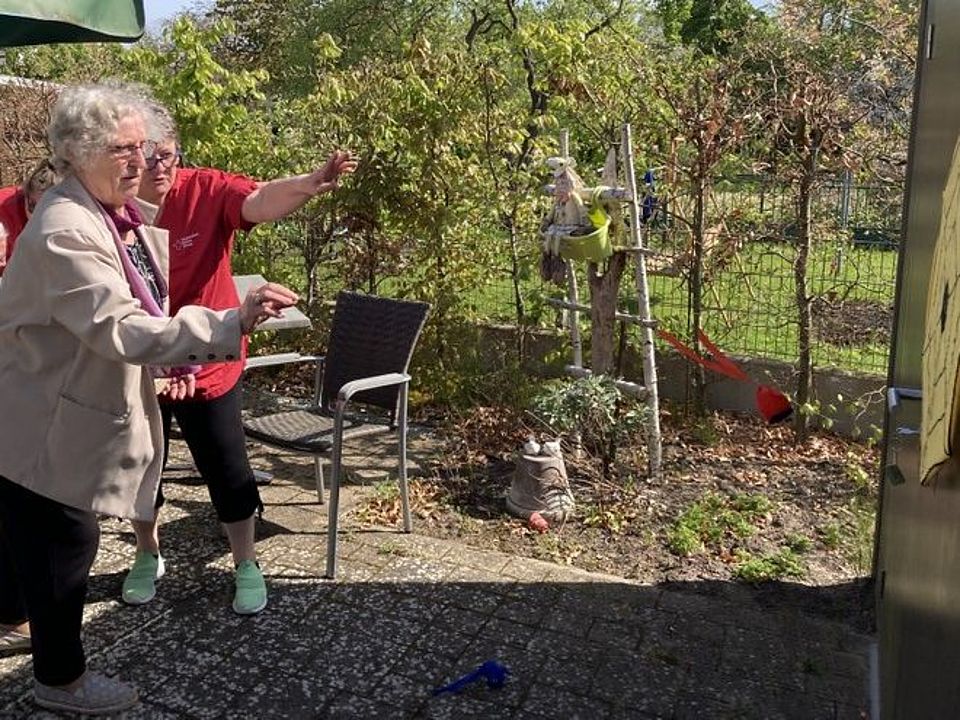 Bild zeigt einen Teil des Außenbereichs wo eine Besucherin der Tagespflege unter Anleitung einer Pflegekraft mit einem Dartspfeil auf eine Dartsscheibe wirft. Diese Aktion ist Teil des alljährlichen Sportfestes