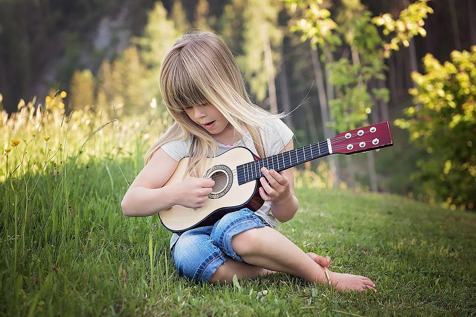 Bild zeigt ein kleines Mädchen auf einer Wiese sitzend mit einer Kindergitarre in der Hand. Im Hintergrund sind leicht verschwommen Bäume zu sehen.
