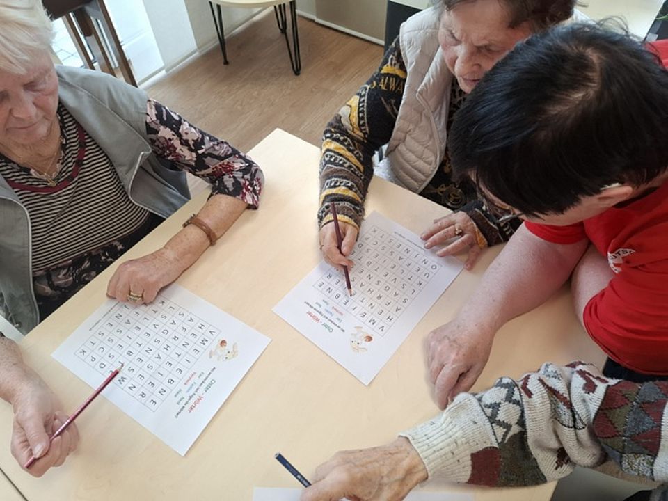Bild zeigt zwei Frauen am Tisch sitzend und jeweils ein Blatt mit Buchstaben in einem Gitter vor sich liegend. beide Frauen halten einen Stift in der rechten Hand und suchen auf dem Blatt Wörter und kreisen diese dann mit dem Stift ein.