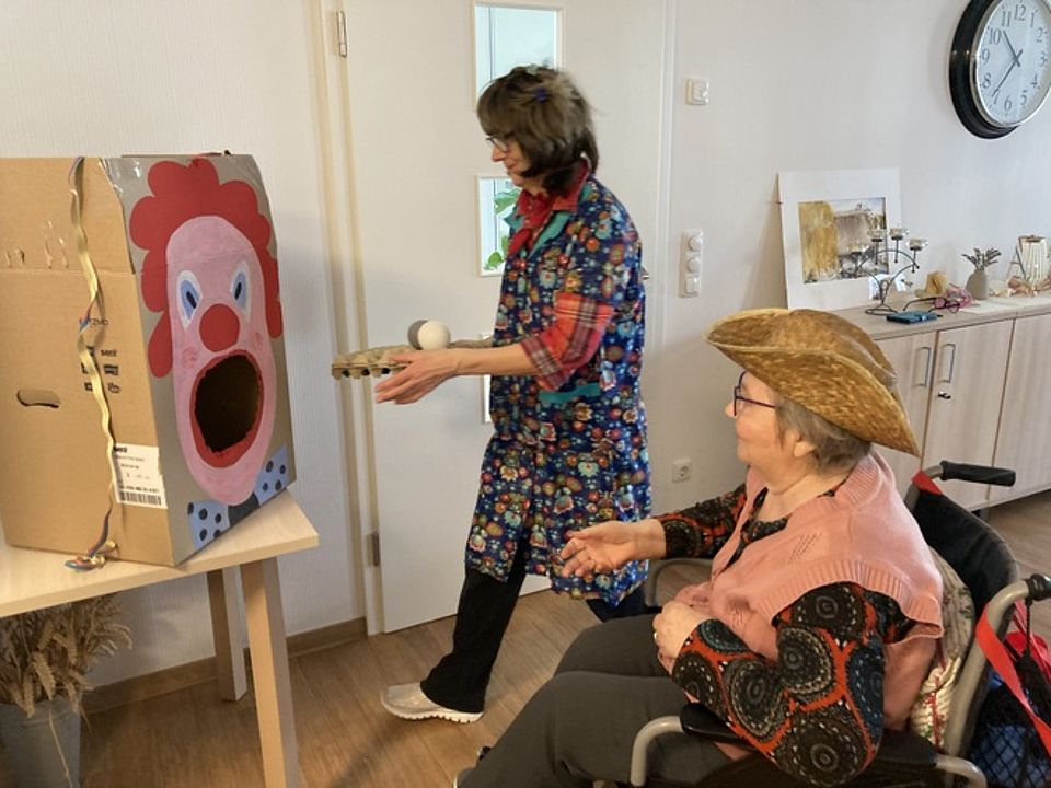 Bild zeigt eine Besucherin der Tagespflege. Sie sitz in ihrem Rollstuhl und hat einen Strohhut auf. Etwas entfernt von ihr steht ein Tisch mit einem lustig bemalten Karton (ein Clownsgesicht). Der Karton hat eine große runde Öffnung (der Mund des Clowns) in den sie versucht Tennisball große Plastikbälle zu werfen.