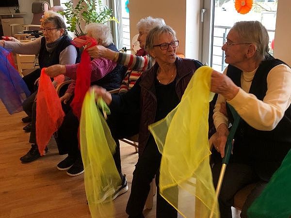 Bild zeigt mehrere Besucherinnen der Tagespflege sitzend in einem Stuhlkreis und bunte Seidentücher nach Musik mit den Händen schwenken