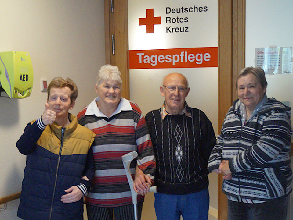 Bild zeigt vier Tagesgäste, 3 Frauen und ein Mann, die Arm in Arm vor der Eingangstür der Tagespflege stehen und gemäß unserem Motto Gemeinsamkeit demonstrieren. An Der Tür im Hintergrund sieht man das Das Rote Kreuz mit dem Schriftzug Deutsches Rotes Kreuz. Darunter ist ein roter Farbbalken in dem in weißen Buchstaben das Wort Tagespflege steht. Die vier Personen schauen mit fröhlich lächelnden Gesichtern in die Kamera.