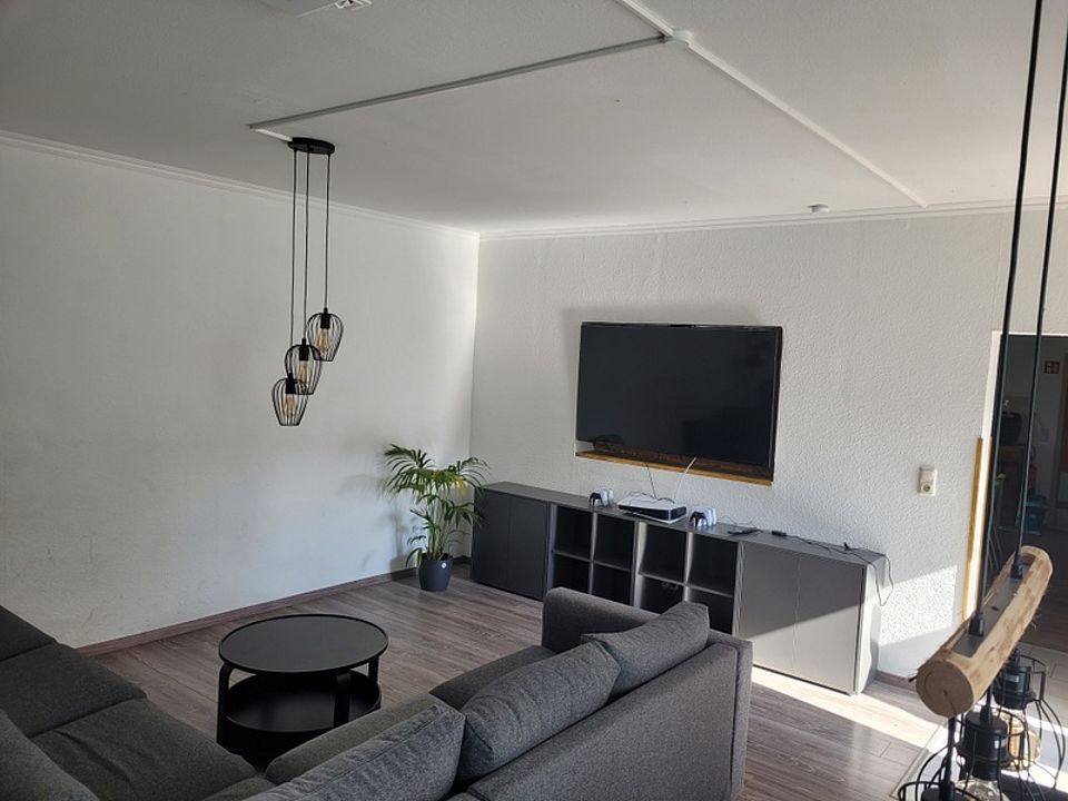 Bild zeigt einen Blick in den großen Wohngemeinschaftsraum mit TV an der Wand, darunter ein Sideboard im Vordergrund eine Sitzlandschaft