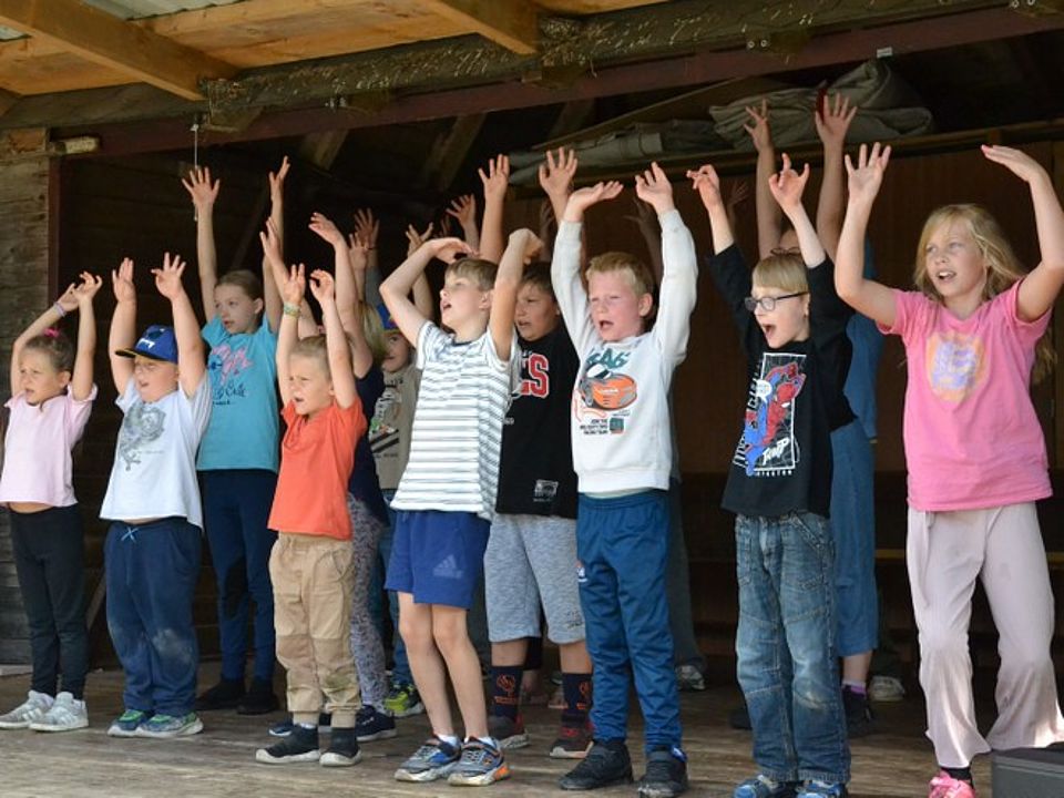 Bild 3 Bild zeigt eine Gruppe von Kindern (12) bei der Programmaufführung. Die Kinder stehen in zwei Reihen und haben die Hände in die Höhe gereckt und singen scheinbar