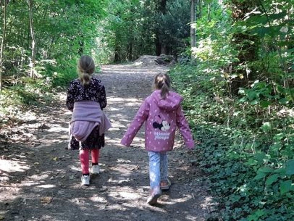 Bild zeigt zwei Mädchen in Rückenansicht, die einen Waldweg spazieren gehen. Sonne scheint durch die Bäume auf den Weg.