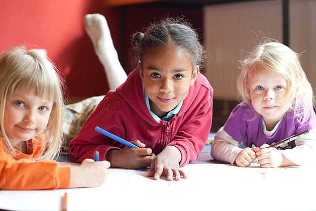 Bild zeigt drei kleine Kinder, welche auf dem Boden liegend mit Buntstiften auf einem Blatt Papier malen