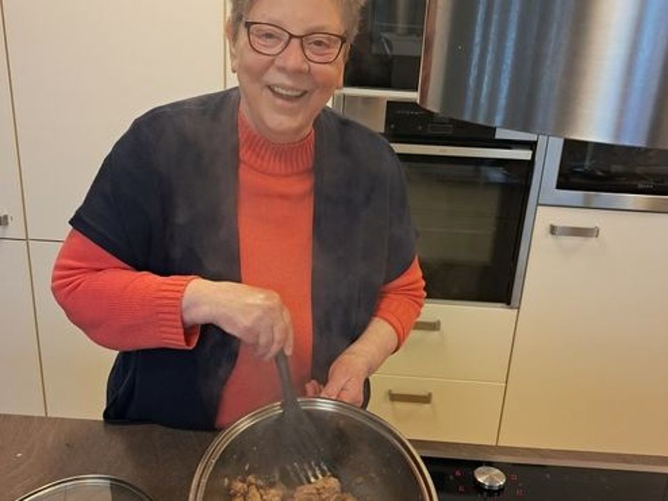 Bild zeigt Frau Monika Boeck am Herd. Sie hebt den den Topf leicht schräg in Richtung Kamera an um so den Inhalt, dass angebratene Fleisch zu zeigen. Frau Boeck ist fröhlich und zeigt ein lachendes Gesicht.