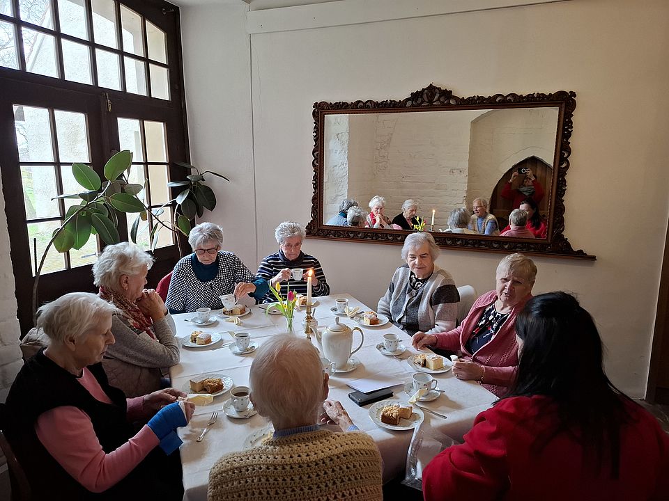 Bild zeigt Eine Gruppe von etwa acht Seniorinnen sitzt gemütlich um einen großen, weiß gedeckten Tisch und genießt bei einem Ausflug eine Kaffeetafel. Auf dem Tisch stehen Tassen, Teller mit Kuchenstücken, eine Teekanne und eine brennende Kerze. Im Hintergrund spiegelt ein großer, dunkler Wandspiegel die Gruppe wider, während links ein großes, dunkles Fenster Tageslicht in den Raum lässt.