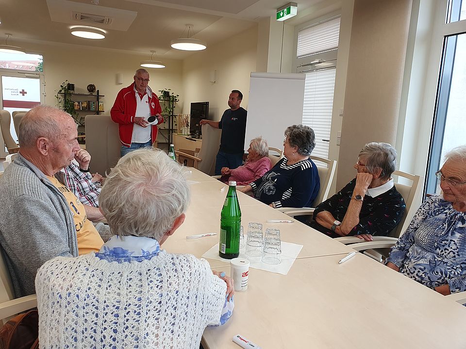Bild zeigt: Ein Vortrag in einem hellen, freundlichen Gemeinschaftsraum. Mehrere Senioren sitzen um einen langen Tisch und hören einem Mitarbeiter des DRK Kreisverbandes Güstrow zu, der das Hausnotrufsystem erklärt. Er hält ein Hausnotrufgerät in der Hand. Auf dem Tisch stehen eine Wasserflasche und Gläser.