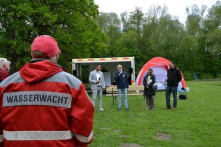 Bild zeigt die 4 in der Bildunterschrift genannten Personen. Sie stehen im Halbkreis auf der Wiese. Präsident Heidenreich hält gerade seine Eröffnungsrede. Im Vordergrund auf der linken Bildseite steht eine Person mit dem Rücken zur Kamera und in DRK Einsatzkleidung und dem Schriftzug Wasserwacht auf dem Rücken.