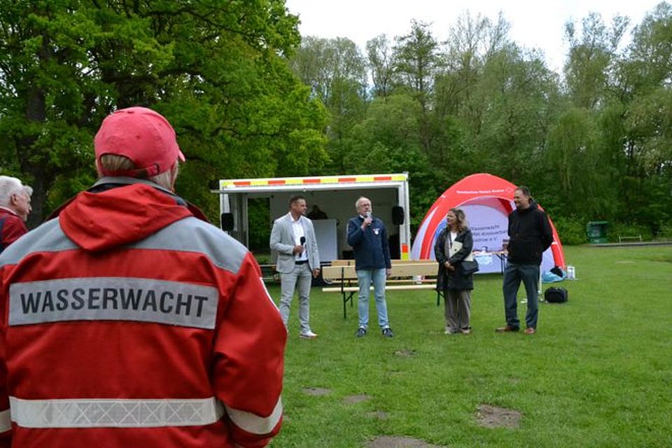 Moderator Christian Schwanbeck Bild zeigt die 4 in der Bildunterschrift genannten Personen. Sie stehen im Halbkreis auf der Wiese. Präsident Heidenreich hält gerade seine Eröffnungsrede. Im Vordergrund auf der linken Bildseite steht eine Person mit dem Rücken zur Kamera und in DRK Einsatzkleidung und dem Schriftzug Wasserwacht auf dem Rücken.