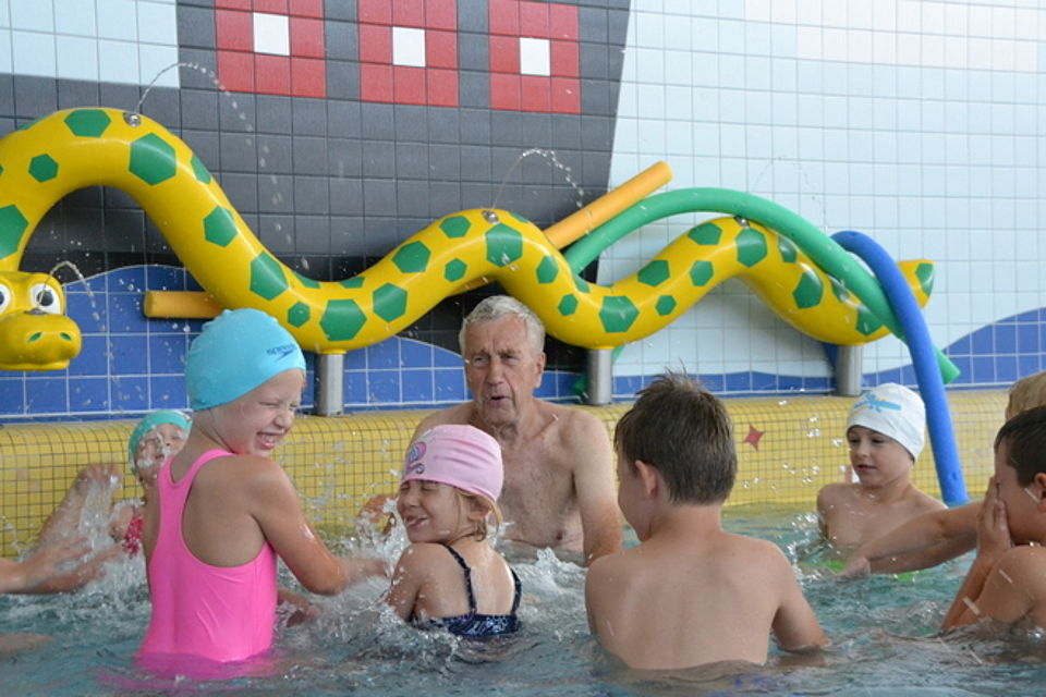Bild zeigt eine Gruppe von 6 Kindern mit Schwimmlehrer Christian Möller im Kinderschwimmbecken zur Eingewöhnung an das Wasser. Allen haben sichtlich Spaß
