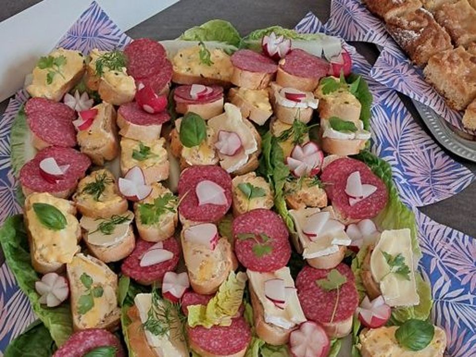 Bild zeigt eine große Platte mit Schnittchen auf einem Salatbett. Die Schnittchen sind mit verschiedenen Wurst- und Käsesorten sowie Salaten belegt und mit Tomatenstückchen, Radieschen, Petersilie und Basilikum blättern garniert.