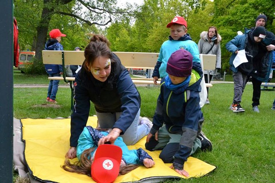 Erste Hilfe Stand Bild zeigt Frau Fehlhaber kniend auf einer gelben Übungsdecke. Vor ihr liegt ein junges Mädchen. An ihr zeigt Frau Fehlhaber die Anwendung der stabilen Seitenlage. Neben ihr kniet ein Junge, der interessiert die Aktion verfolgt.